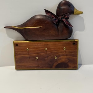 Peut inclure: Un canard en bois brun foncé avec un bec et des accents dorés, portant un nœud à motifs, est posé sur un porte-clés en bois avec des crochets en laiton. Le porte-clés est rectangulaire.