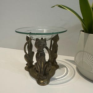 Puede incluir: Un cuenco decorativo de vidrio sostenido por una base de metal color bronce con tres figuras de gato. Los gatos están sentados, con un gato en el centro y dos a los lados. El cuenco es transparente y redondo, y la base tiene un diseño ornamentado.