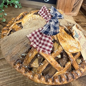 May include: A decorative apple pie with a lattice crust, adorned with red and blue plaid bows and a burlap wrap. The pie is placed on a wooden surface, with a tag that reads "Apple Pie". The pie is a rustic, country-style decoration.