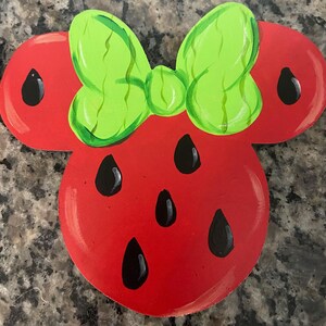 May include: A red wooden decoration in the shape of a mouse head with a green bow. The mouse head is painted to resemble a watermelon, with black teardrop-shaped seeds.