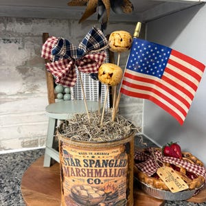 May include: A decorative arrangement featuring a vintage-style tin can labeled "Star Spangled Marshmallow Co." with marshmallow skewers, fabric bows, and a small American flag. A miniature strawberry pie sits nearby on a wooden tray.
