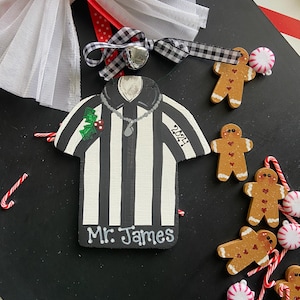 May include: A black and white striped referee ornament with the text "Mr. James" and "2021" on it. The ornament is surrounded by gingerbread men and candy canes on a black background.