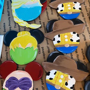 May include: Five painted wooden Mickey Mouse ears featuring different Disney characters. The characters include Elsa, Woody, Tinkerbell, Ariel, and another Woody. The ears are painted in bright colors and have a whimsical design.