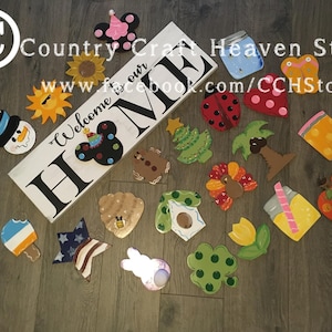 May include: A collection of wooden painted ornaments featuring various themes, including a snowman, a sun, a Mickey Mouse head, a Christmas tree, a palm tree, a turkey, a beehive, a birdhouse, a tulip, a four-leaf clover, a popsicle, a star, a bunny, a mason jar, a butterfly, a ladybug, and an apple. The ornaments are arranged on a wooden surface. The text "Welcome to our Home" is painted on a white wooden sign.