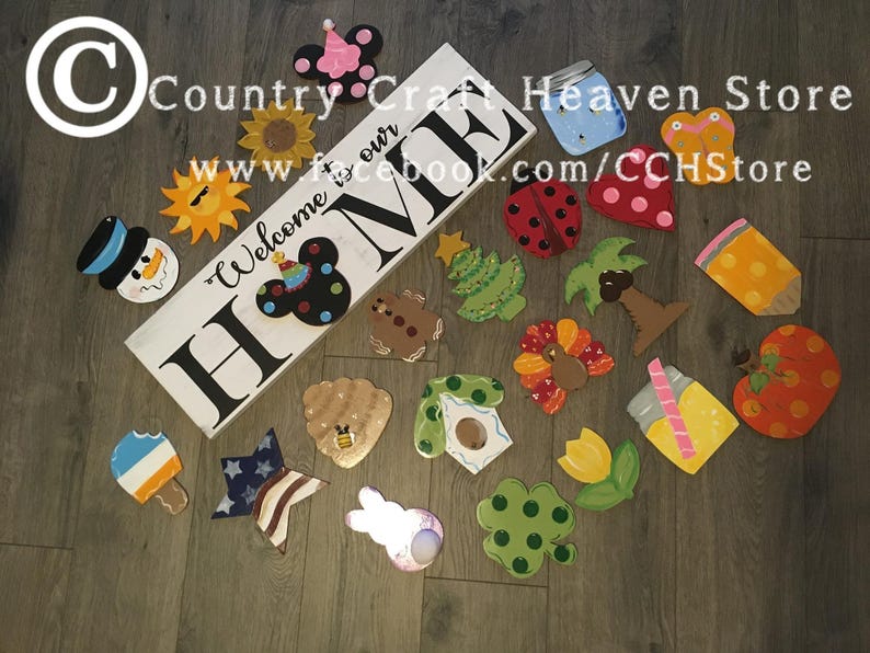 May include: A collection of wooden painted ornaments featuring various themes, including a snowman, a sun, a Mickey Mouse head, a Christmas tree, a palm tree, a turkey, a beehive, a birdhouse, a tulip, a four-leaf clover, a popsicle, a star, a bunny, a mason jar, a butterfly, a ladybug, and an apple. The ornaments are arranged on a wooden surface. The text "Welcome to our Home" is painted on a white wooden sign.