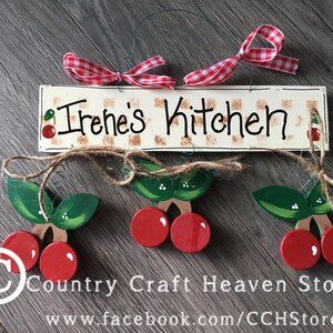 May include: A wooden sign with the text "Irene's Kitchen" painted in brown lettering on a white background. The sign is decorated with red and green painted cherries and a red and white checkered ribbon. The sign is hanging from a twine string with three additional painted cherry ornaments.