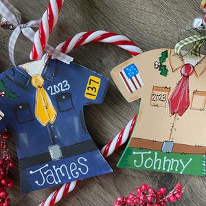 May include: Two wooden ornaments shaped like shirts, one blue with a yellow tie and the name "James" and the other tan with a red tie and the name "Johnny". Both shirts have a patch with the American flag and a number on them.
