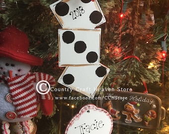 May include: A white and black dice ornament with the year 2018 on one side. The ornament is hanging on a Christmas tree with red and white gingham ribbon. A white oval ornament with the name Tracie is hanging on the tree below the dice ornament.