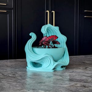 May include: A light turquoise planter featuring a sculptural design that mimics the appearance of crashing ocean waves. The bowl-shaped container holds a small plant with dark red and green leaves, resting on a matching integrated base.