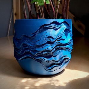 Puede incluir: Una maceta azul, impresa en 3D, con un diseño en forma de ola. La maceta está llena de una planta verde con tallos de color rojo oscuro. La maceta está sobre una superficie de madera.