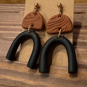 Peut inclure: Paire de boucles d'oreilles faites à la main avec un design bicolore. La partie supérieure est une forme semi-circulaire marron avec une texture de grain de bois, reliée à un fond noir incurvé en forme d'arche. Des accents métalliques dorés sont utilisés pour les clous d'oreilles et la quincaillerie de connexion.