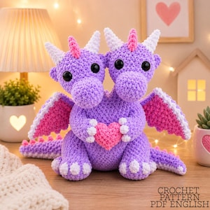 Pode incluir: Um brinquedo de pelúcia de dragão de duas cabeças feito em crochê roxo com chifres brancos, detalhes rosa nas asas e na cabeça, e um coração rosa segurado nas patas dianteiras. O texto CROCHET PATTERN PDF ENGLISH está impresso no canto inferior.