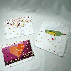 May include: Three fabric envelopes with envelope-style flaps. Two envelopes feature a white background with small red, pink, and gold heart patterns. One envelope displays a large, geometric heart design in shades of orange, pink, and dark purple.