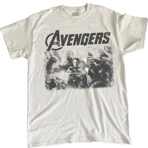 May include: White t-shirt featuring a distressed graphic of the Avengers team. The shirt displays the word "Avengers" in a bold, black font above a grayscale image of the superhero team. A crew neck and short sleeves complete the design.