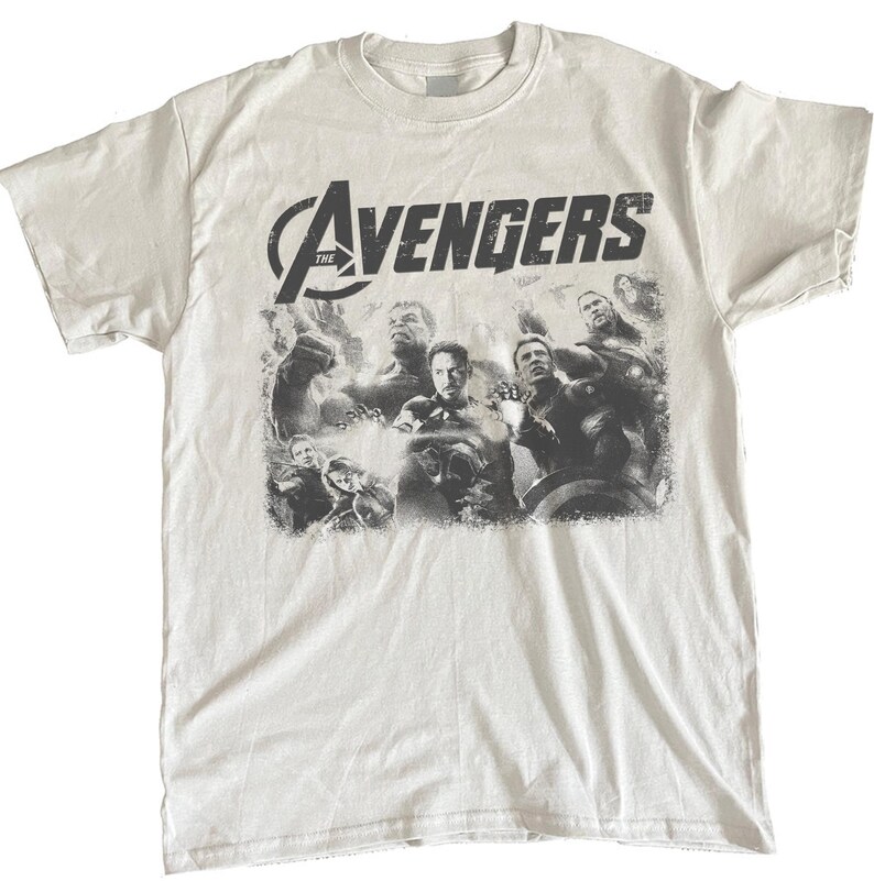 May include: White t-shirt featuring a distressed graphic of the Avengers team. The shirt displays the word "Avengers" in a bold, black font above a grayscale image of the superhero team. A crew neck and short sleeves complete the design.