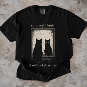 May include: A black short-sleeve t-shirt featuring a graphic of a cat sitting in front of a mirror with its reflection. The text "i do not think" is printed above the mirror and "therefore i do not am" is printed below the mirror in white.