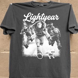 May include: Dark gray t-shirt featuring the word "Lightyear" in white script above a graphic of Buzz Lightyear and other characters running. A white cat is also running in the graphic.