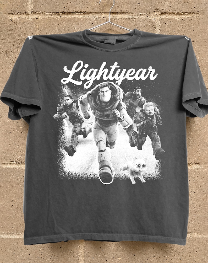 May include: Dark gray t-shirt featuring the word "Lightyear" in white script above a graphic of Buzz Lightyear and other characters running. A white cat is also running in the graphic.