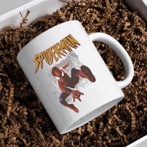 Puede incluir: Una taza de cer&aacute;mica blanca con un gr&aacute;fico del personaje Spider-Man en una pose de salto din&aacute;mica. Sobre el personaje, la palabra SPIDERMAN est&aacute; impresa en una fuente dorada estilizada y dentada. La taza tiene un asa curva y acabado liso.