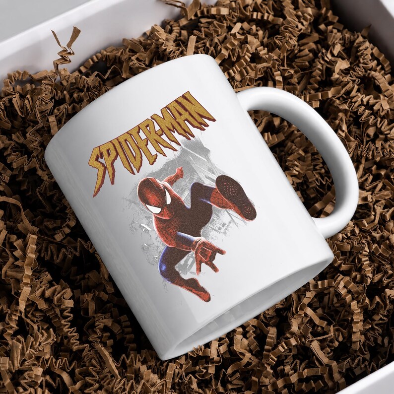 Puede incluir: Una taza de cer&aacute;mica blanca con un gr&aacute;fico del personaje Spider-Man en una pose de salto din&aacute;mica. Sobre el personaje, la palabra SPIDERMAN est&aacute; impresa en una fuente dorada estilizada y dentada. La taza tiene un asa curva y acabado liso.