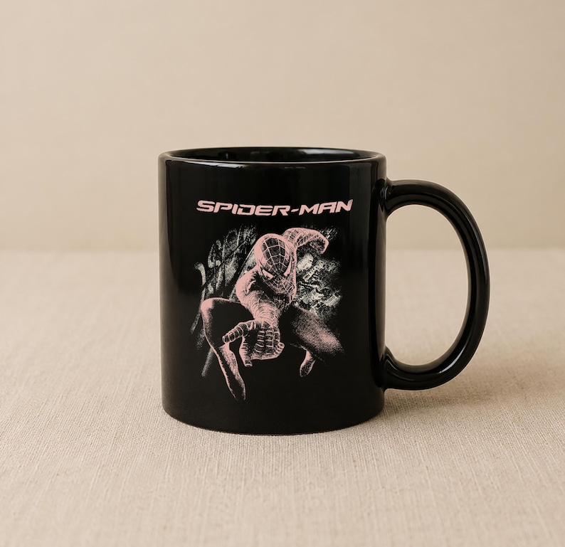 Puede incluir: Una taza de cer&aacute;mica negra con un gr&aacute;fico rosa claro del personaje Spider-Man en pose agachada. El texto "SPIDER-MAN" est&aacute; impreso en una fuente rosa claro sobre la ilustraci&oacute;n del personaje. La taza tiene un acabado liso y un asa.