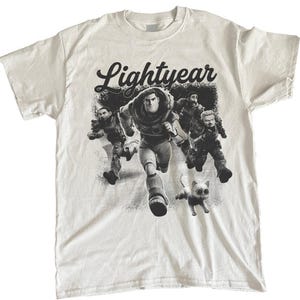 May include: White t-shirt featuring a black and white graphic of characters from the movie "Lightyear." The design includes the word "Lightyear" in a stylized font above a running Buzz Lightyear and other characters, with a small cat.