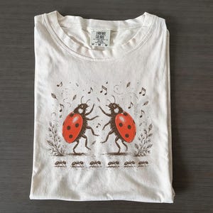 May include: An off-white short-sleeve t-shirt featuring a graphic of two ladybirds dancing, surrounded by musical notes and small leaves. Below the ladybirds is a line of six ants. The shirt has a Comfort Colors brand tag at the neck.