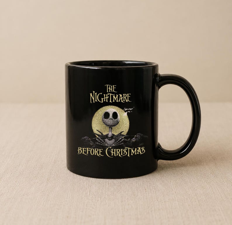 May include: A black ceramic mug featuring a graphic of a skeletal character in a pinstripe suit standing before a large yellow moon. The text "THE NIGHTMARE BEFORE CHRISTMAS" is printed in a stylized yellow font above and below the character illustration.