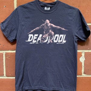 Puede incluir: Camiseta azul marino con un dise&ntilde;o de Deadpool en una pose din&aacute;mica, sosteniendo dos espadas. La palabra "DEADPOOL" est&aacute; impresa en blanco. La camiseta est&aacute; hecha de un material suave.