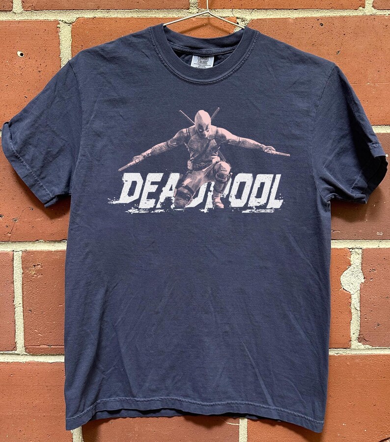 Puede incluir: Camiseta azul marino con un dise&ntilde;o de Deadpool en una pose din&aacute;mica, sosteniendo dos espadas. La palabra "DEADPOOL" est&aacute; impresa en blanco. La camiseta est&aacute; hecha de un material suave.