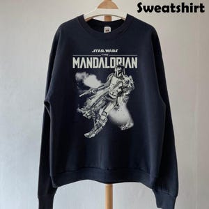 Op de afbeelding: Zwart sweatshirt met een afbeelding van The Mandalorian uit Star Wars. Het ontwerp bevat de tekst "STAR WARS THE MANDALORIAN" in het wit, en een afbeelding van het personage. Het sweatshirt hangt aan een houten hanger.