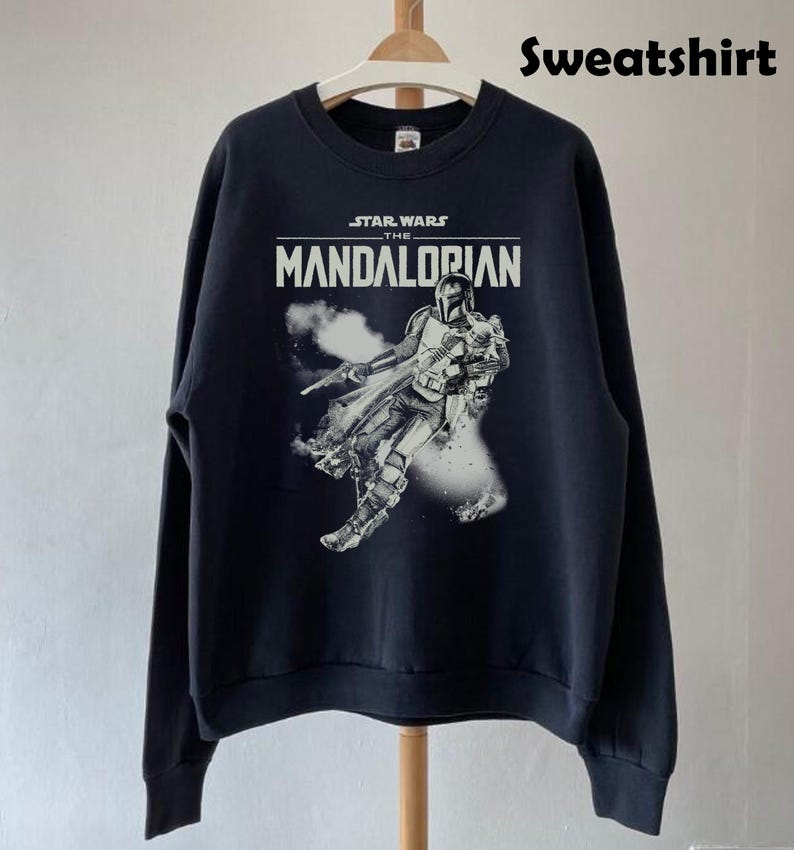Op de afbeelding: Zwart sweatshirt met een afbeelding van The Mandalorian uit Star Wars. Het ontwerp bevat de tekst "STAR WARS THE MANDALORIAN" in het wit, en een afbeelding van het personage. Het sweatshirt hangt aan een houten hanger.