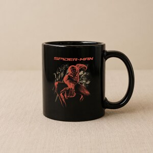 May include: A black ceramic mug featuring a red graphic of the character Spider-Man in a crouching pose. Above the character, the text SPIDER-MAN is printed in a bold red font. The mug has a smooth, glossy finish and a curved handle on the right side.