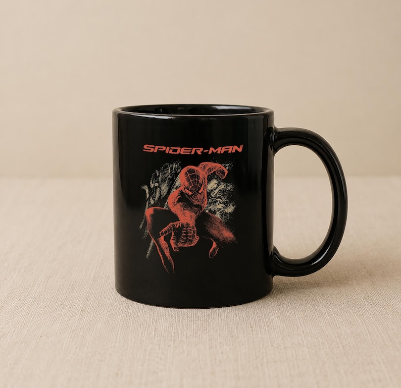 May include: A black ceramic mug featuring a red graphic of the character Spider-Man in a crouching pose. Above the character, the text SPIDER-MAN is printed in a bold red font. The mug has a smooth, glossy finish and a curved handle on the right side.