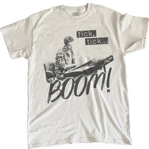 May include: White t-shirt featuring a black and white graphic of a person playing a keyboard, with the words "tick, tick" above and "BOOM!" below. The design is printed on a short-sleeved, crew neck t-shirt.