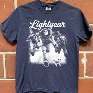 May include: Navy blue t-shirt featuring the word "Lightyear" in white script above a graphic of Buzz Lightyear and other characters. The graphic is in black and white, with a white cat at the bottom.