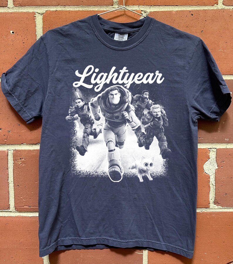 May include: Navy blue t-shirt featuring the word "Lightyear" in white script above a graphic of Buzz Lightyear and other characters. The graphic is in black and white, with a white cat at the bottom.