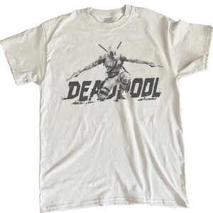 Puede incluir: Camiseta blanca con un gr&aacute;fico en blanco y negro del personaje de Deadpool arrodillado con armas. La palabra "DEADPOOL" est&aacute; impresa en una fuente desgastada debajo del personaje.