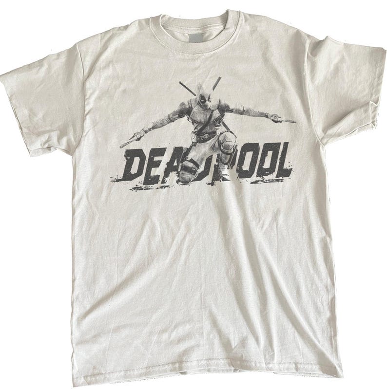Puede incluir: Camiseta blanca con un gr&aacute;fico en blanco y negro del personaje de Deadpool arrodillado con armas. La palabra "DEADPOOL" est&aacute; impresa en una fuente desgastada debajo del personaje.