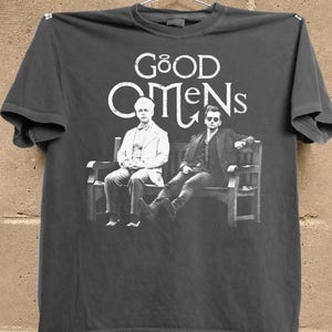 May include: Dark gray t-shirt featuring the text "GOOD OMENS" above a black and white image of two men seated on a bench. The image is a graphic design, likely for fans of the show.