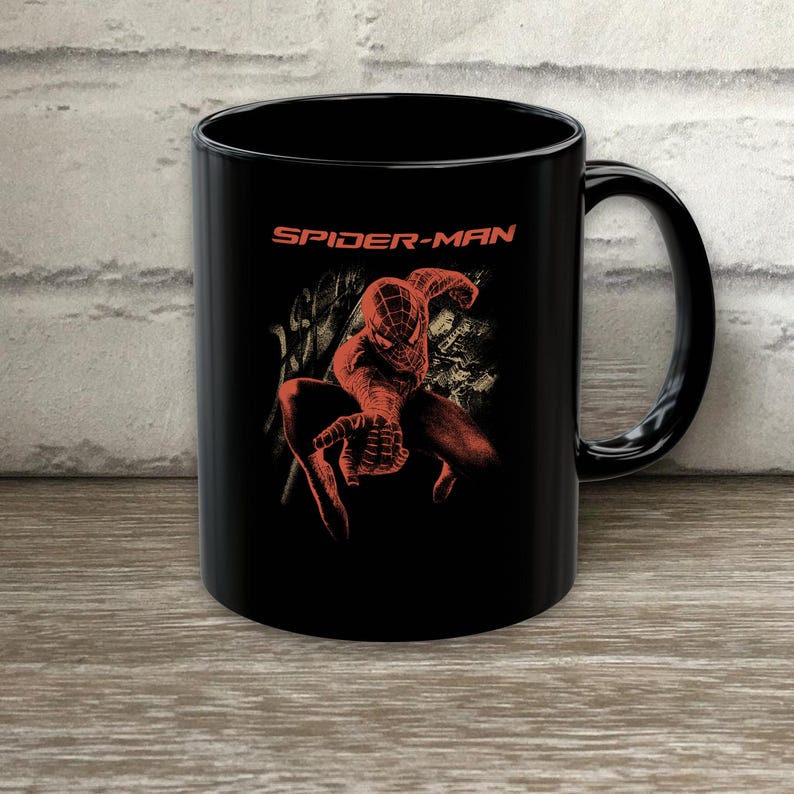 May include: A black ceramic mug featuring a red graphic of the character Spider-Man in a dynamic pose with one hand extended forward. The text SPIDER-MAN is printed in orange above the character illustration on the front of the mug.