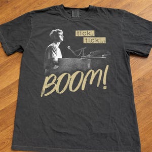 May include: Dark gray t-shirt featuring a black and white image of a person at a piano with a microphone. The shirt has the words "tick, tick..." and "BOOM!" in a tan color.