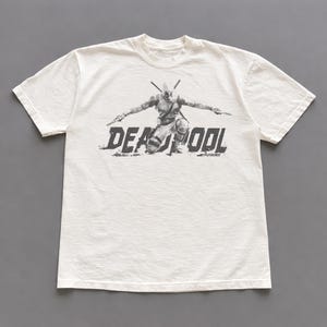 Puede incluir: Camiseta blanquecina con un gr&aacute;fico en blanco y negro de Deadpool arrodillado con armas. La palabra "DEADPOOL" est&aacute; impresa en una fuente desgastada debajo del personaje. La camiseta tiene mangas cortas y cuello redondo.
