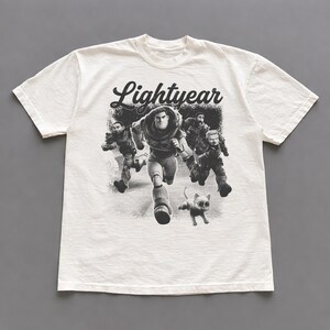 May include: Off-white t-shirt featuring a black and white graphic of characters running, with the word "Lightyear" in a stylized font. The design includes a cat and several figures in space suits.