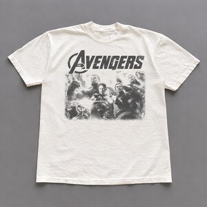 May include: Off-white t-shirt featuring a distressed graphic of the Avengers team. The word "AVENGERS" is printed across the top in a distressed font. The shirt is made of a soft, comfortable material.