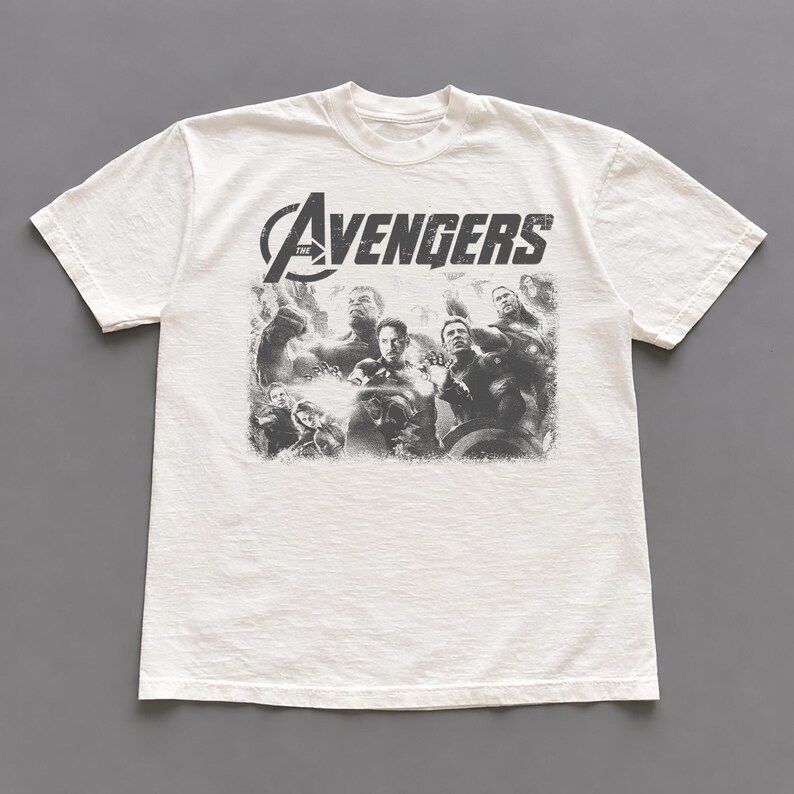 May include: Off-white t-shirt featuring a distressed graphic of the Avengers team. The word "AVENGERS" is printed across the top in a distressed font. The shirt is made of a soft, comfortable material.