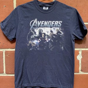 May include: Navy blue t-shirt featuring a graphic of The Avengers team. The Avengers logo is in white and silver. The shirt is hanging on a metal hanger against a brick wall.
