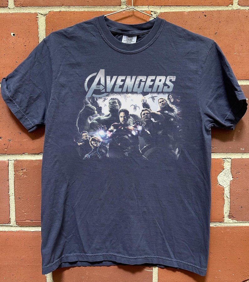 May include: Navy blue t-shirt featuring a graphic of The Avengers team. The Avengers logo is in white and silver. The shirt is hanging on a metal hanger against a brick wall.