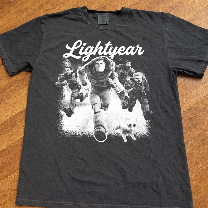 May include: Dark gray t-shirt featuring a graphic from the movie "Lightyear." The design shows Buzz Lightyear running with other characters and a cat. The word "Lightyear" is written in white script across the top.