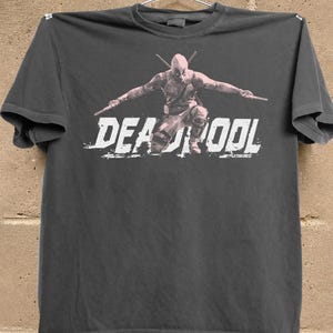 Puede incluir: Camiseta gris oscuro con un gr&aacute;fico de Deadpool en posici&oacute;n de combate, sosteniendo dos espadas. La palabra "DEADPOOL" est&aacute; impresa en blanco, con letras desgastadas en la parte inferior de la imagen.